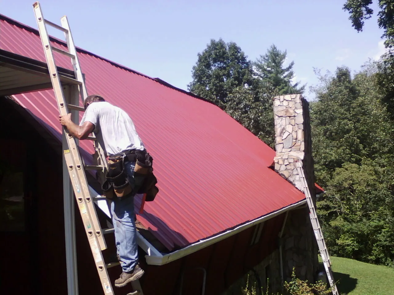 Roofer on a ladder beside a red metal roof and stone chimney — Soffit & Fascia Repair by Roofing Brothers in Olympia Fields